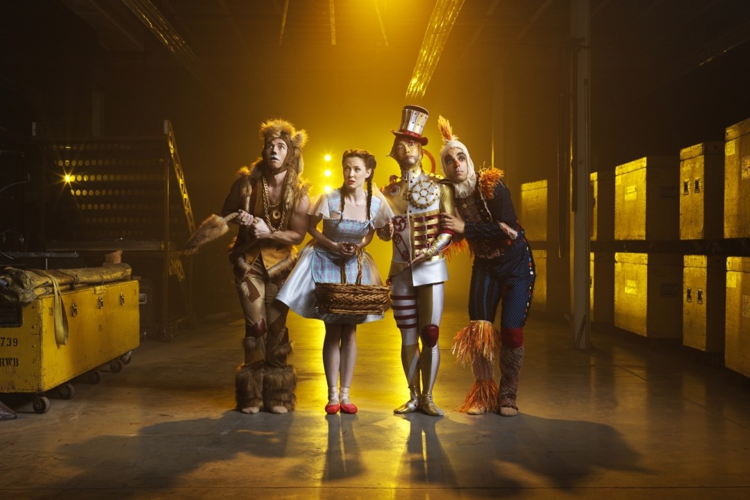 Man in lion costume, woman in blue dress and red shoes, man in tin man costume, and man in scarecrow costume standing next to each other in front of a yellow background.