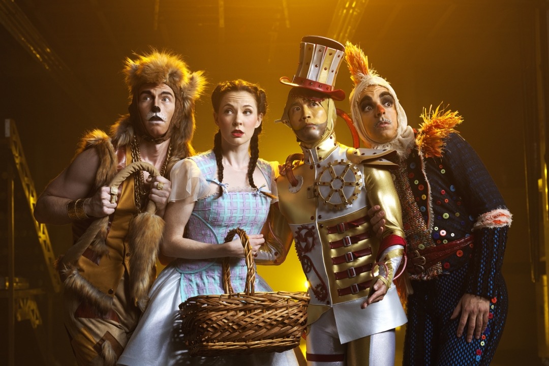 Man in lion costume, woman in blue dress and red shoes, man in tin man costume, and man in scarecrow costume standing next to each other in front of a yellow background.