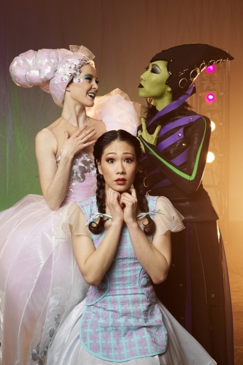 Woman in a pink dress and pink hair next to a green woman in a black dress with a black hat behind a woman in a blue dress sitting in front of a brown, lit up background.