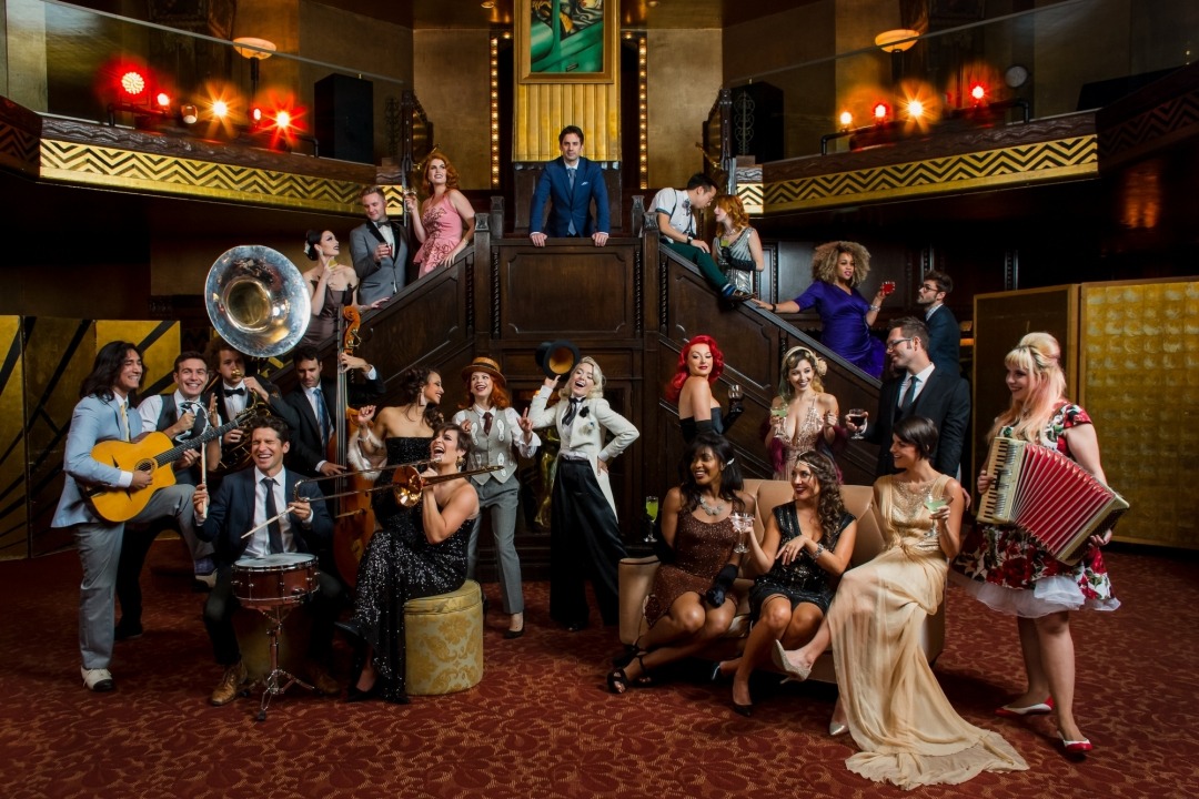22 people in a 1920's styled room with brown, burgandy, and gold colors all variously standing and sitting. Some with instruments, some with whine glasses, all dressed in various 1920's costumes.