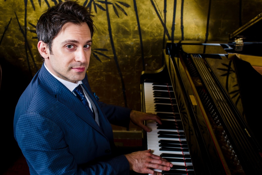 White man dressed in a blue suit with a blue and white striped tie, facing the camera as he plays the piano.