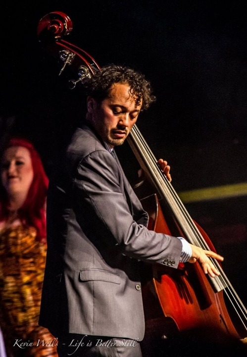Man with black, curly hair, is wearing a grey suit and playing the bass instrument. White women with bright red hair, wearing a yellow and black dress, is standing behind the man.