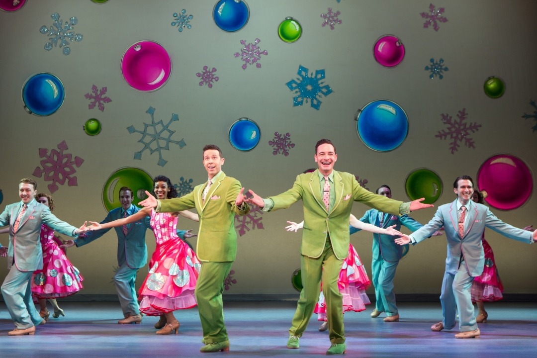 Two men in green suits dancing in front of a row of people in gray suits and red dresses dancing in front of a yellow background with ornaments and snowflakes on it