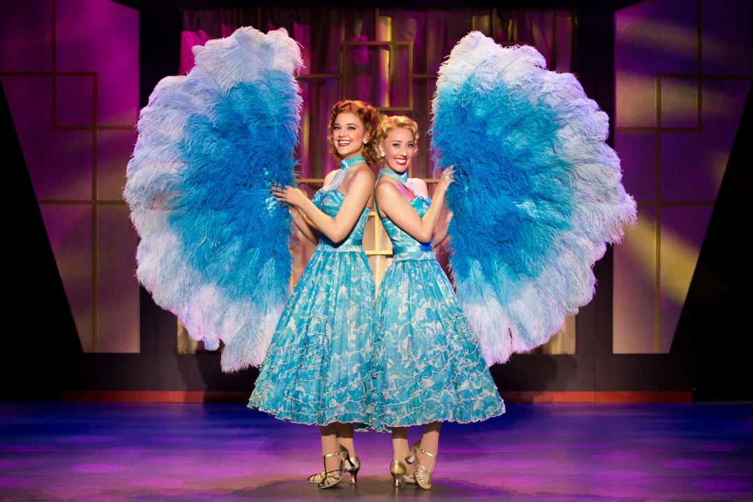 Two women in blue dresses holding big blue and white fans in front of a purple background