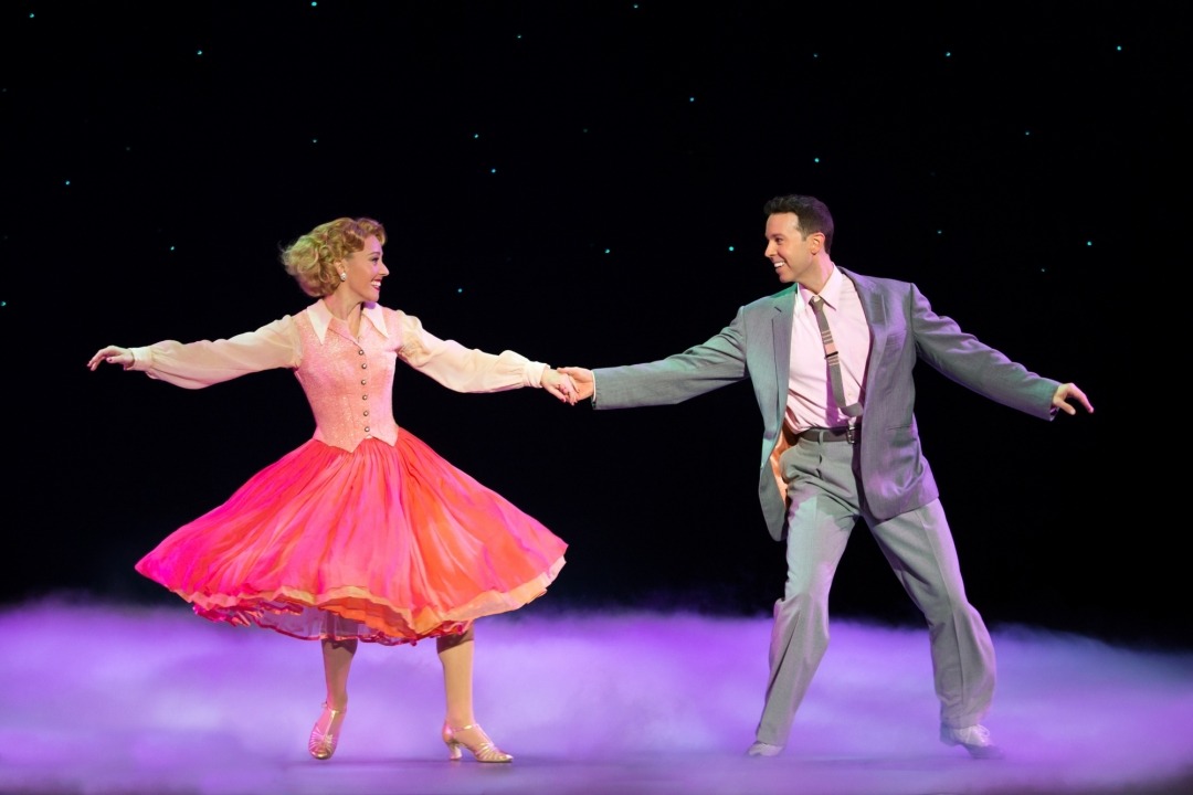 A woman in a pink dress and a man in a gray suit dancing in front of a black background
