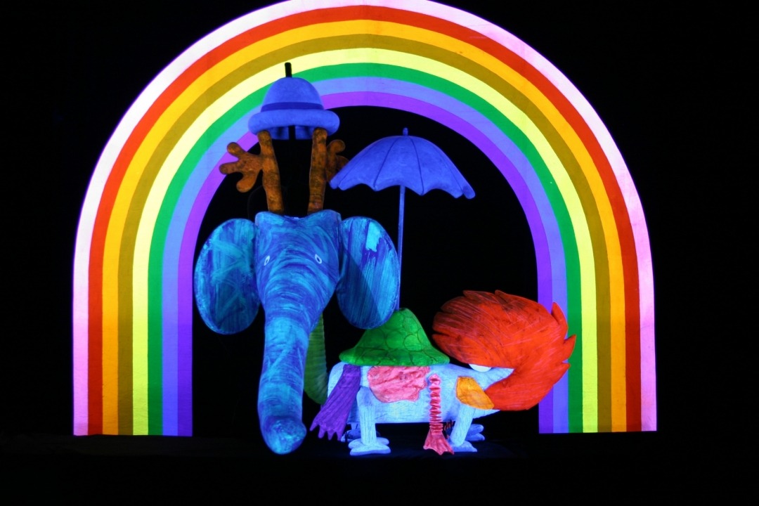 An imaginative animal puppet with an elephant head, a fox tail, a turtle back, and antlers wearing a hat and carrying an umbrella under a vibrant rainbow