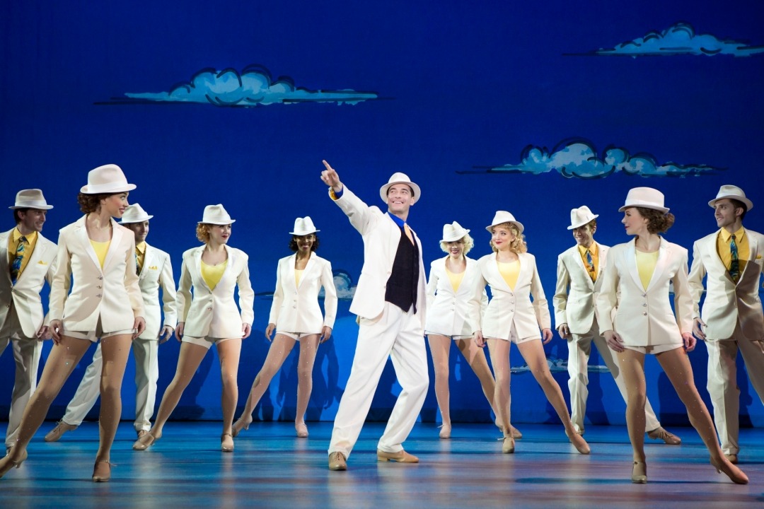 Man in white suit pointing upwards in front of women in white suits standing in front of a blue sky background