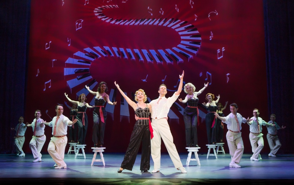 Man in white suit and woman in black outfit standing with their arms up in front of a line of people in the same outfits in front of a red background with piano keys