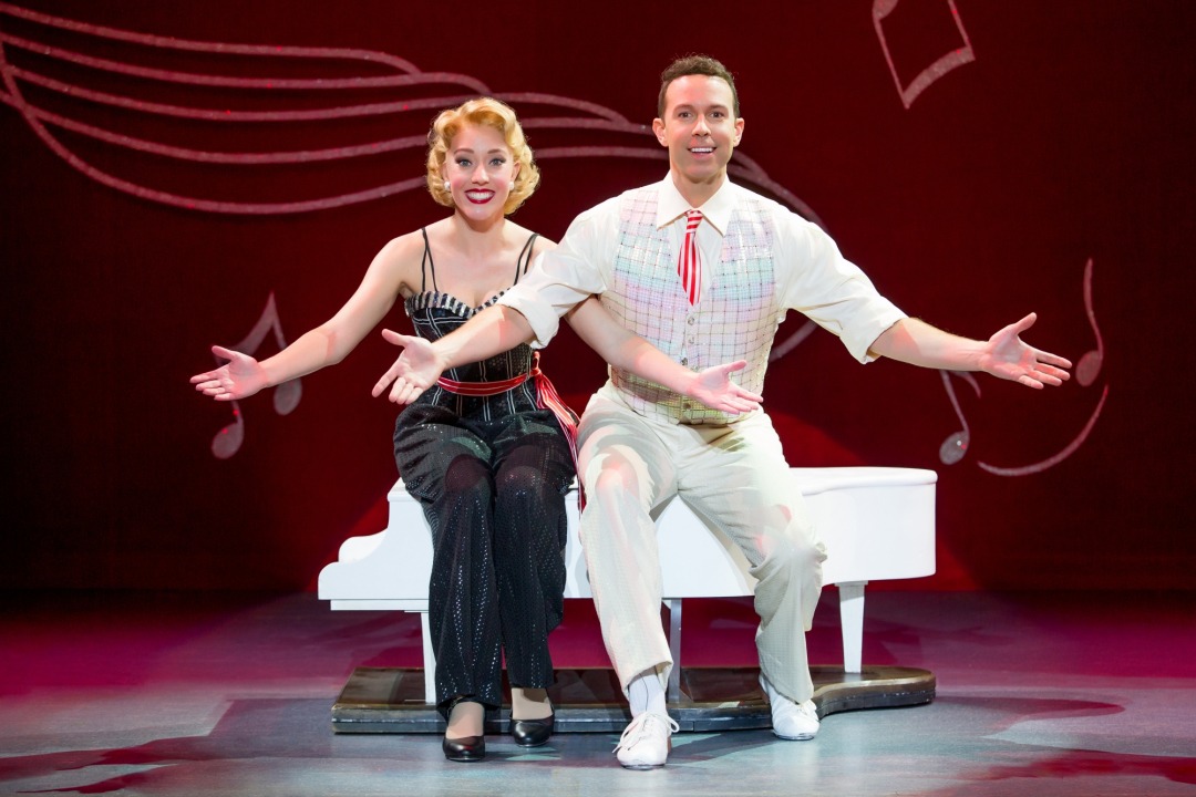 Man in white outfit and woman in black outfit sitting on a small white piano with arms presented forward
