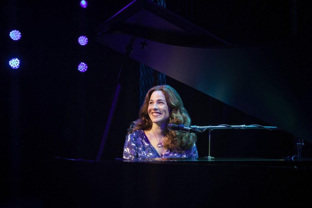 Woman in purple dress sitting behind a grand piano and microphone smiling