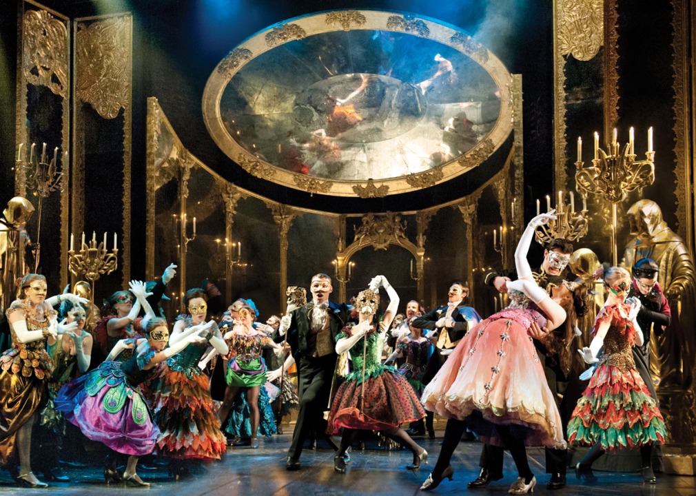 Dancers in masks and colorful gowns in front of a gold ballroom background