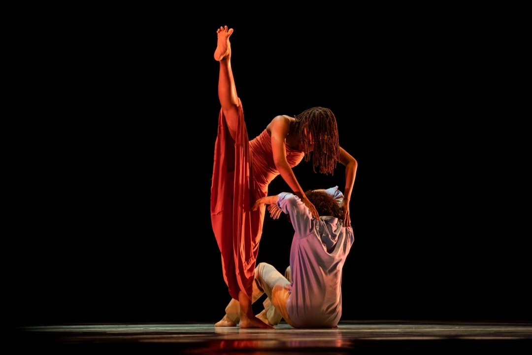 Man sitting on the ground with a girl pressing both hands on his shoulders with her leg in the air.