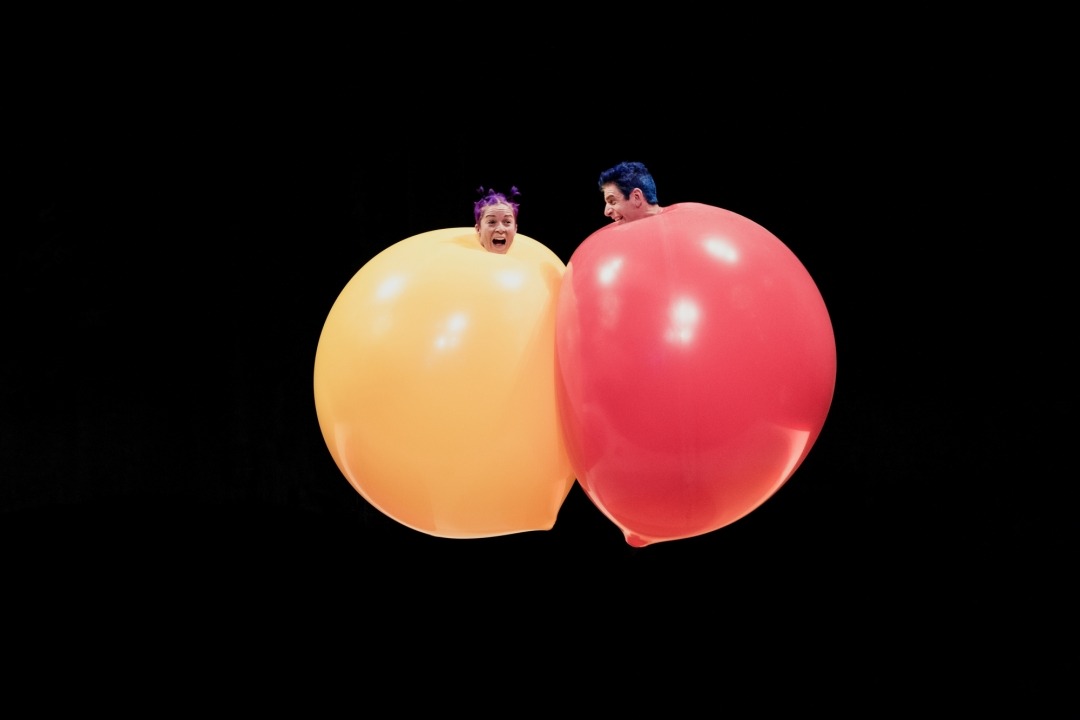 Woman with her body in a giant yellow balloon and a man with his body in a giant red balloon in front of a black background.