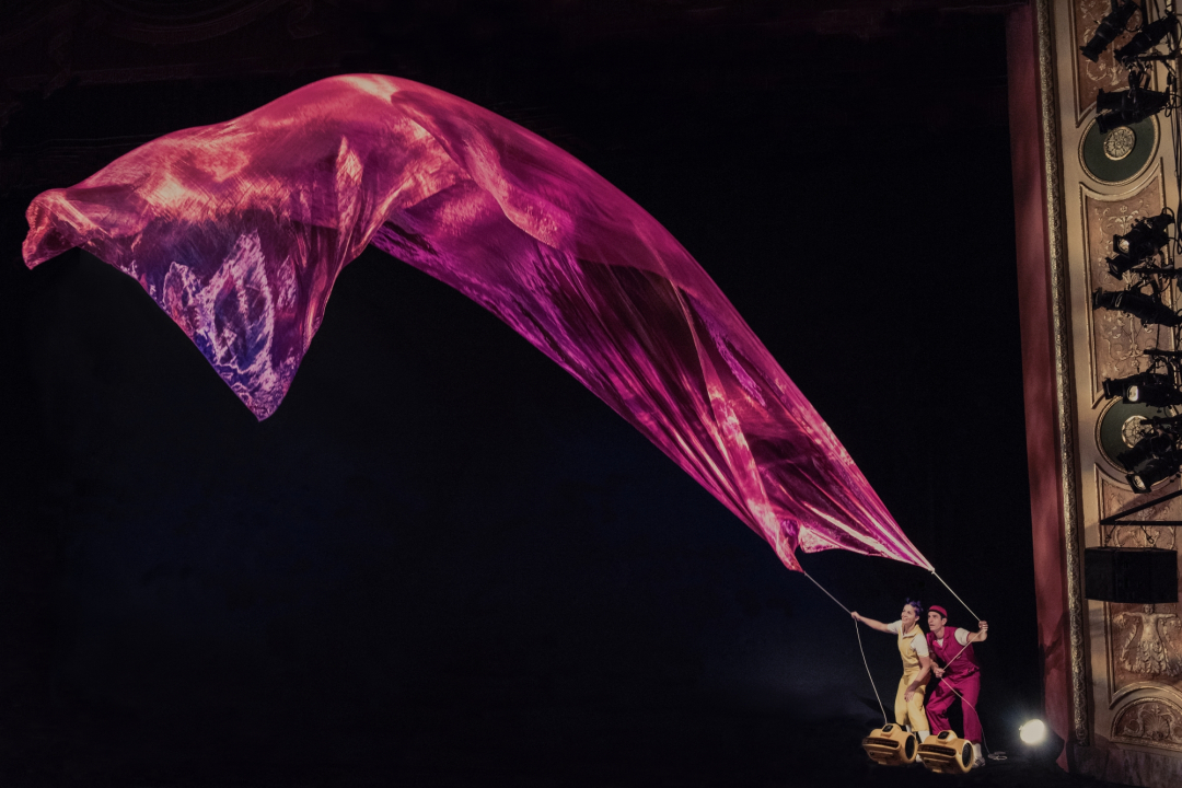 A woman in yellow and a man in red holding a string connected to a red piece of sheer fabric that's floating in front of a black background.
