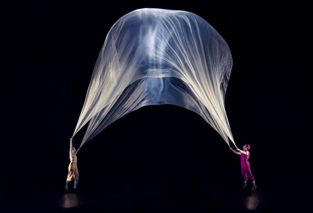 Woman in a yellow outfit and man in a red outfit holding sheer white fabric that's floating in front of a black background.