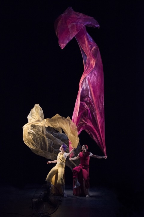 Woman in a yellow outfit holding a sheer yellow floating fabric and man in a red outfit holding a sheer red floating fabric in front of a black background.