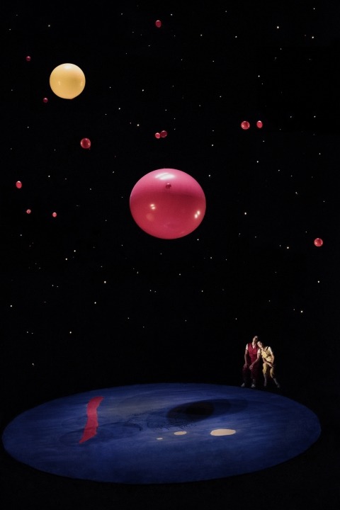 A woman in yellow and a man in red sitting at the corner of a stage looking up at floating red and yellow balloons in front of a black background.