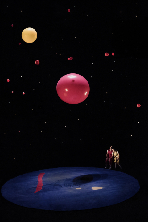 A woman in yellow and a man in red sitting at the corner of a stage looking up at floating red and yellow balloons in front of a black background.