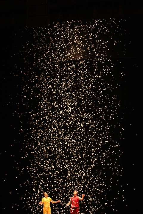 Woman in a yellow outfit and man in a red outfit with yellow confetti falling from the ceiling in front of a black background.