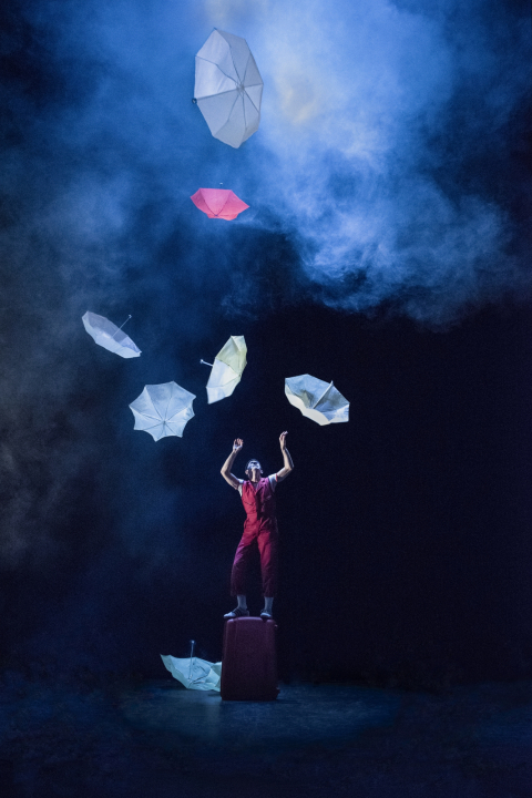 Man in red outfit standing on a red suitcase with seven umbrellas floating around him in front of a black and blue, smokey background.