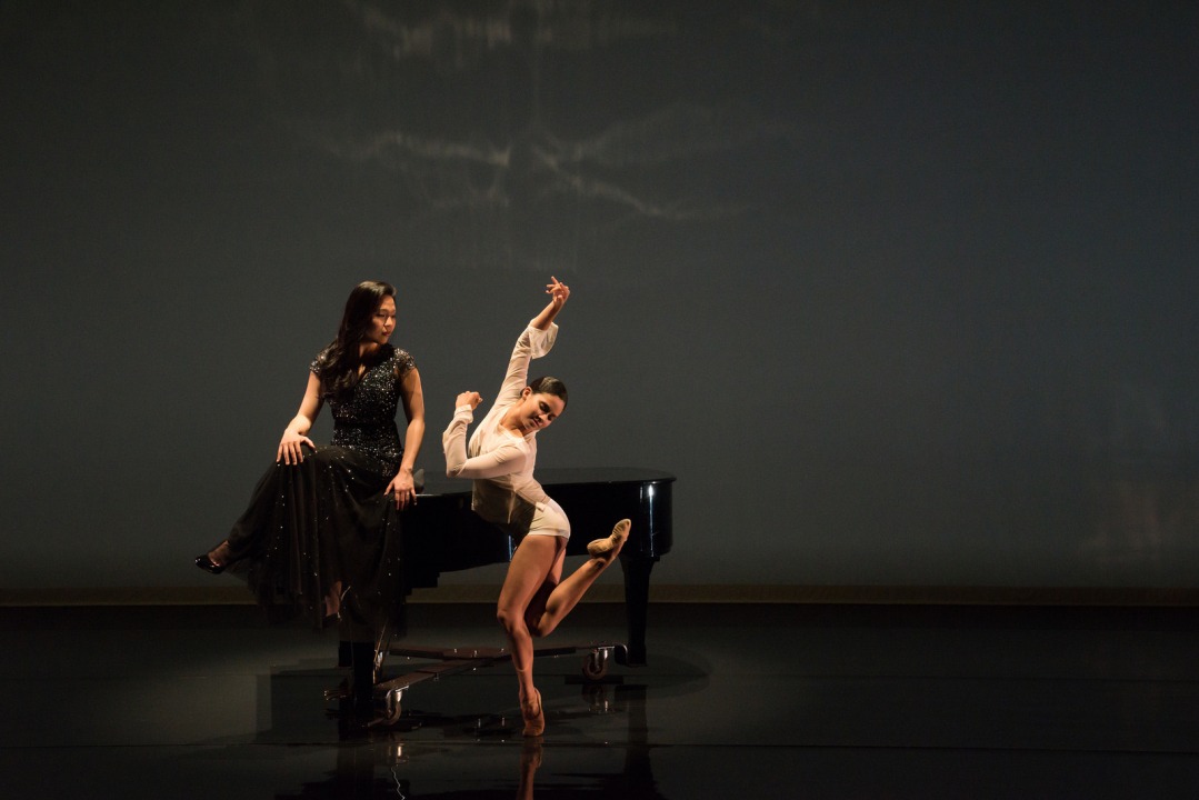 Women wearing a long black dress is sitting on the piano body facing another women who is dancing next to the piano and she dressed in white.