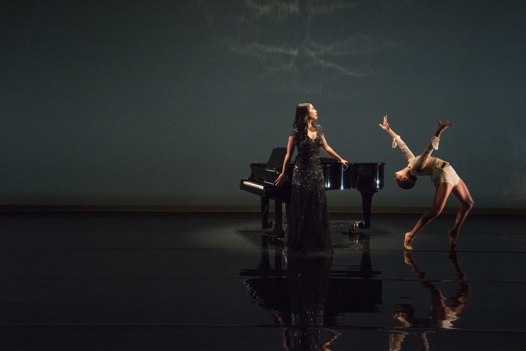 Woman wearing a long black dress standing in the middle of the frame, with a grand piano behind her. A women dressed in white dances to the women's left.