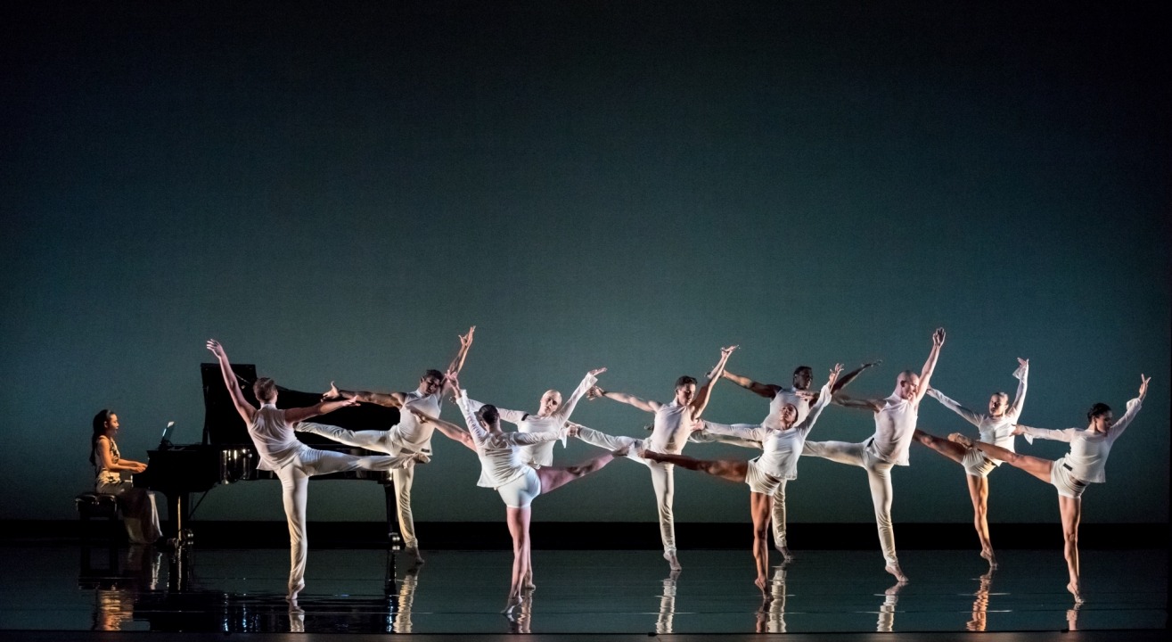 Nine women dressed in white dancing in front of a black background, with a piano and player in the back left corner