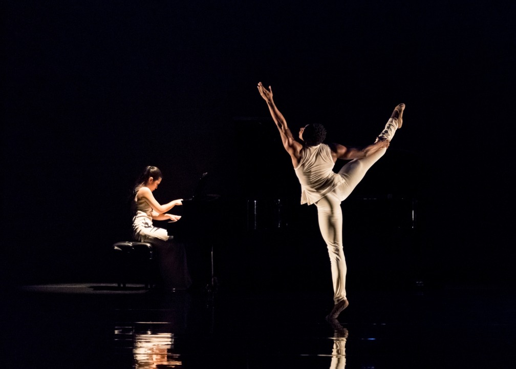 A man dressed in white dancing while facing the back, black background. Asian American woman playing on the piano in the background to the left.