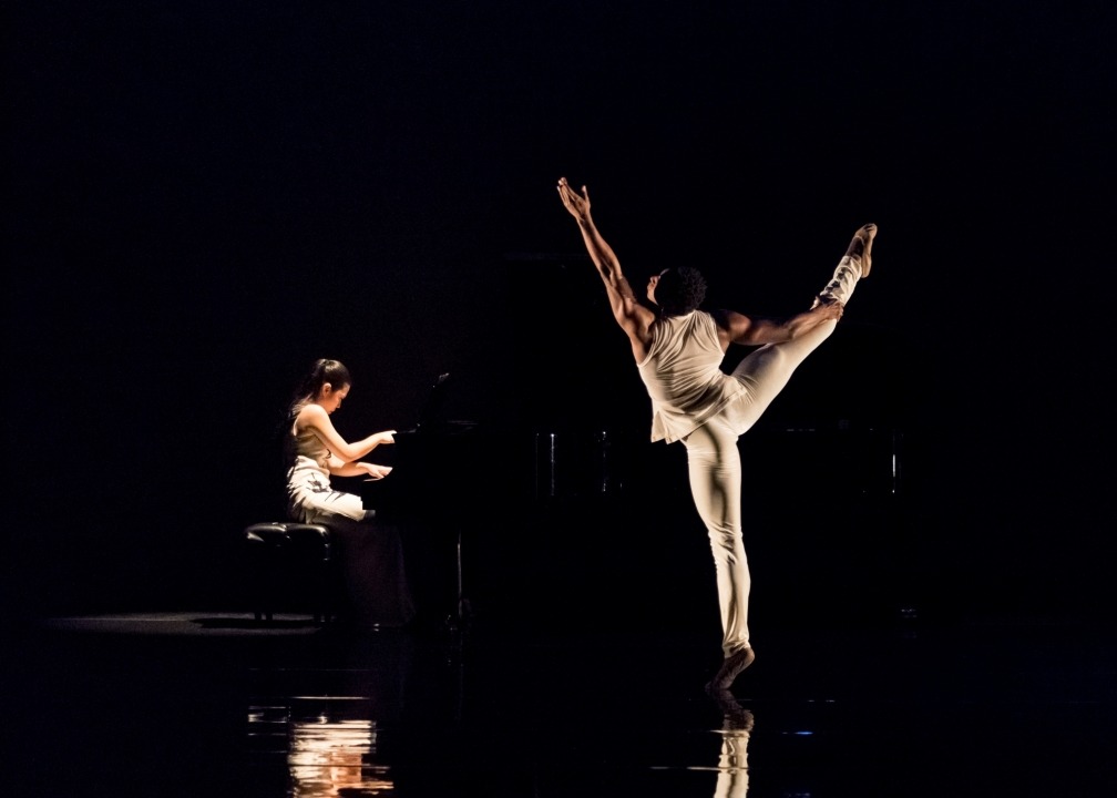 A man dressed in white dancing while facing the back, black background. Asian American woman playing on the piano in the background to the left.