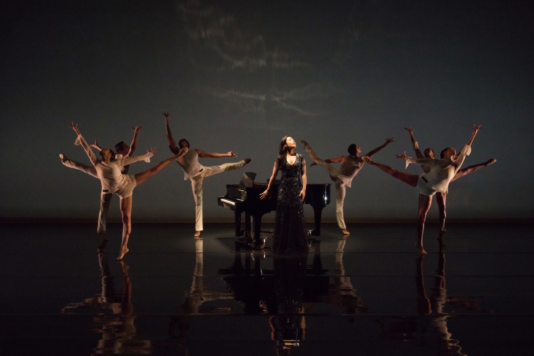 Women wearing a long black dress is standing in the middle of the stage, with a grand piano behind her. The women in the black dress is surrounded by six dancers in white - three to her right and three to her left. The background is black.