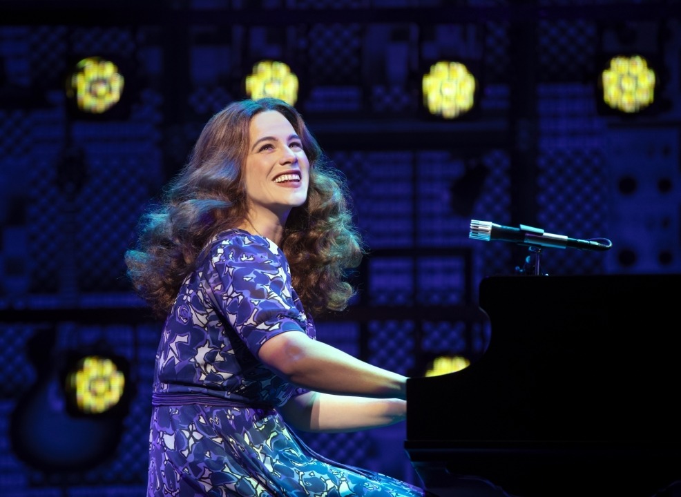 Woman in purple dress sitting at a grand piano singing in front of lights