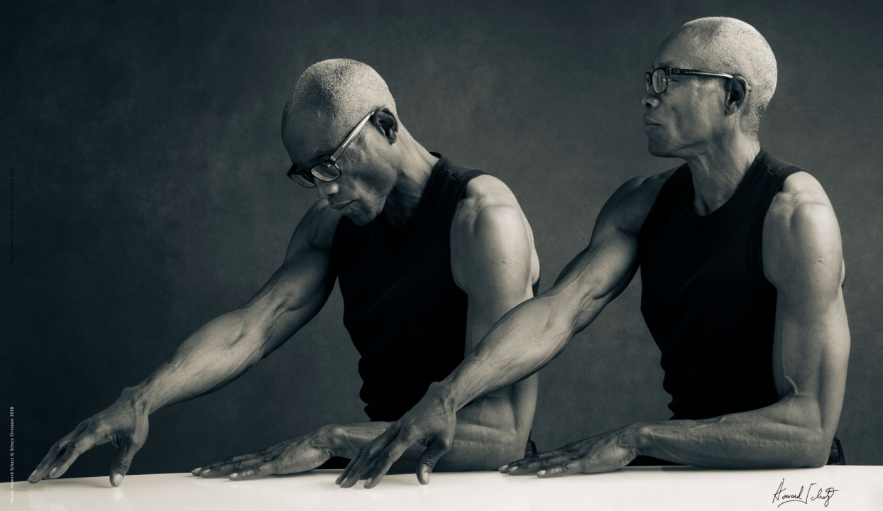 Two black and white images of the same man at a table in a black shirt and glasses in front of a grey background