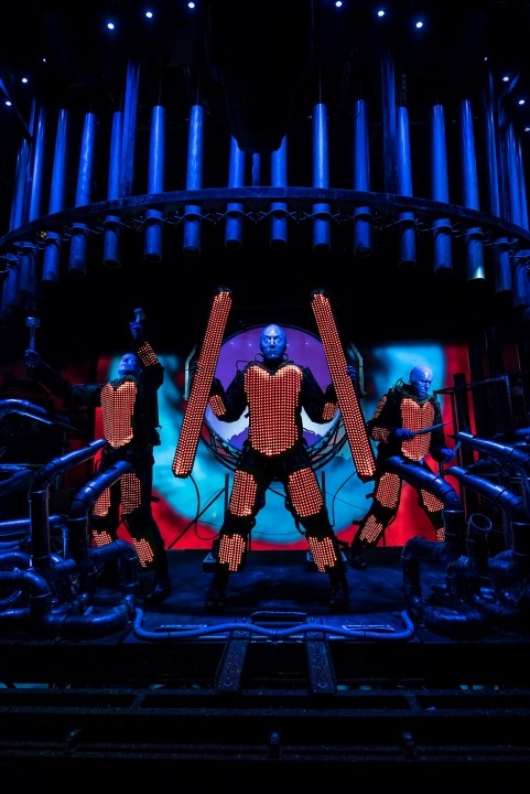 3 blue bald men wearing outfits with red lights on them in front of a blue background