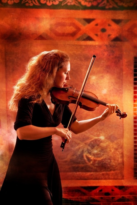 A woman in a black dress playing a violin in front of an orange background.