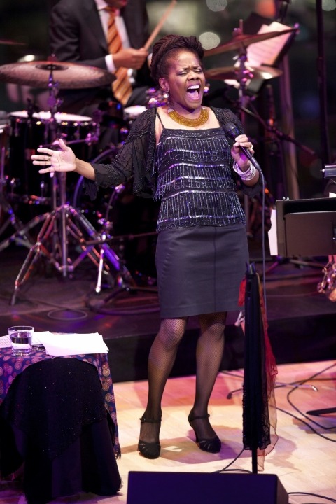 African American women, wearing a black sparkly shirt and a black skirt, is singing strongly into the microphone. Drummer wearing a yellow, white, and purple tie is playing in the background