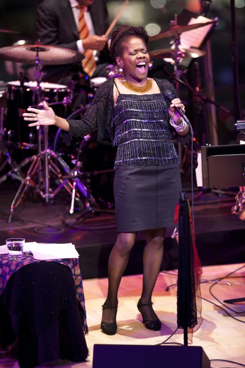 African American women, wearing a black sparkly shirt and a black skirt, is singing strongly into the microphone. Drummer wearing a yellow, white, and purple tie is playing in the background