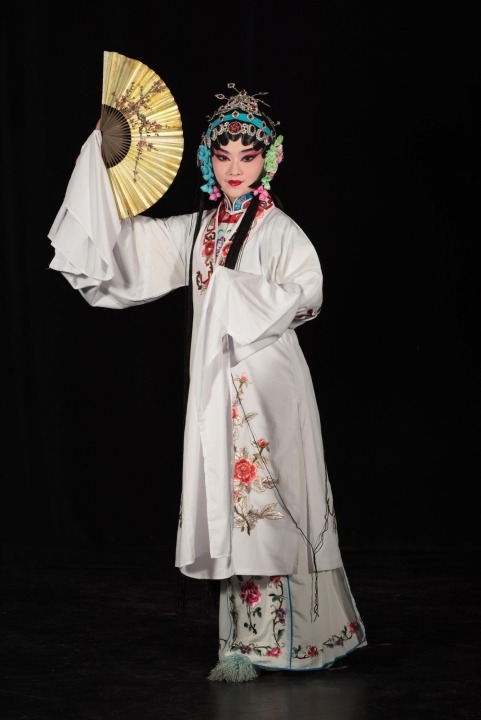 A Chinese woman wearing a white robe with flowers on it and a gold and blue headpiece stands holding a gold fan vertically near her face in front of a black background.
