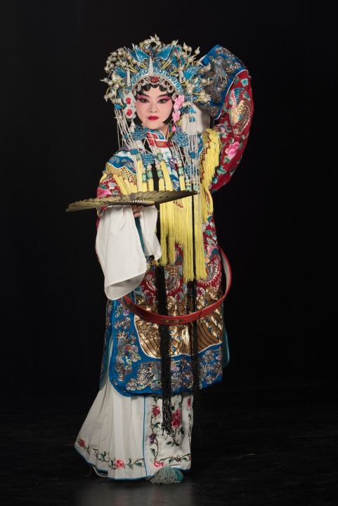 A Chinese woman wearing a headdress and robe made of ornate red, white, yellow, and blue fabric stands holding out a gold fan in front of a black background.