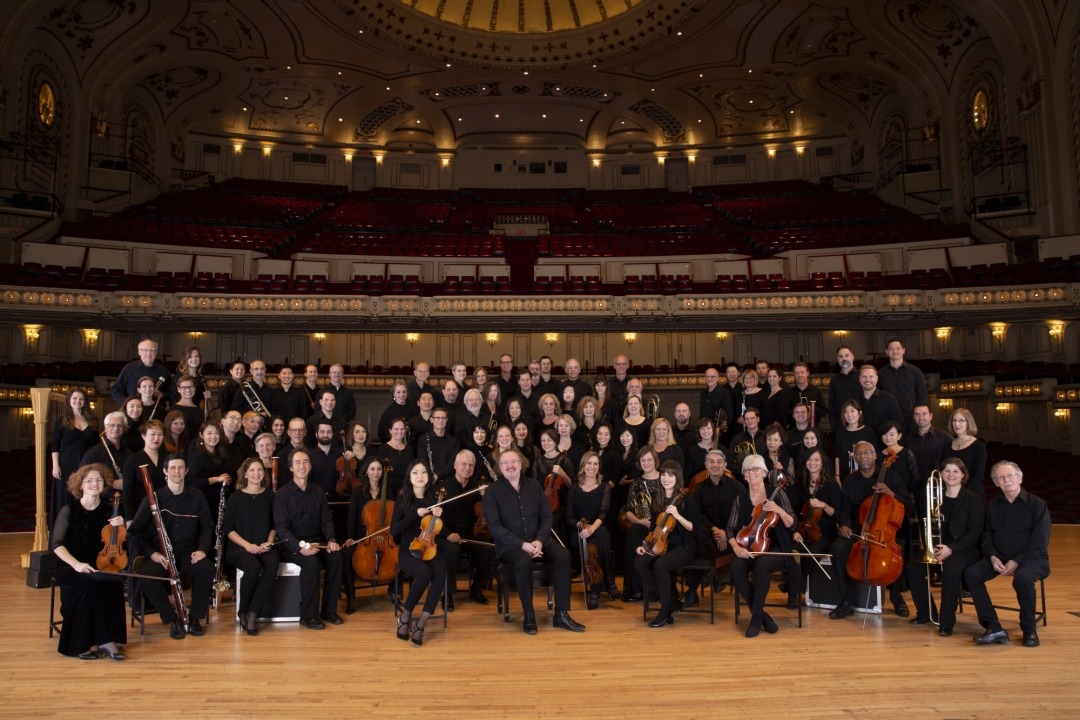 The St. Louis Symphony all dressed in black holding their instruments smiling at the camera in front of the seats of an auditorium.