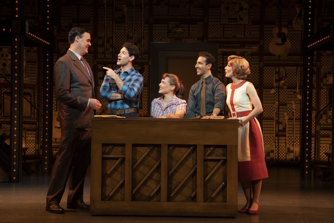Three men and two women sitting behind a piano talking in front of a brown background