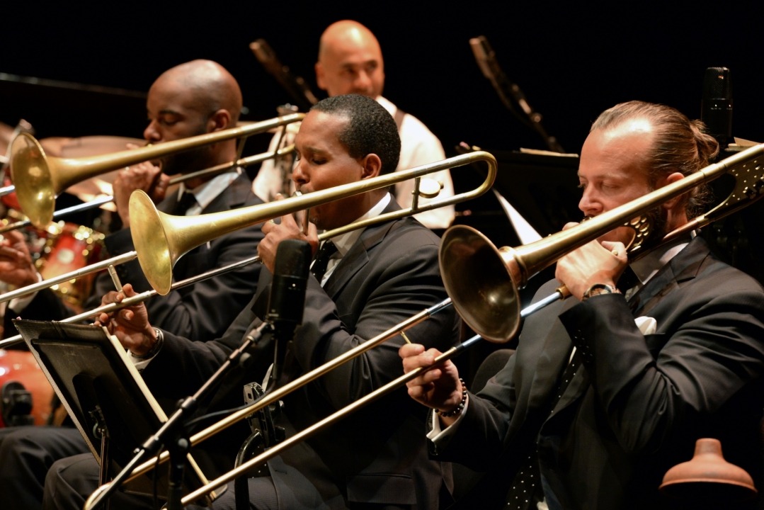 Three men, wearing black suits with black and white dotted ties, are playing the trombone sitted in a line. Drum player is visible in the background.