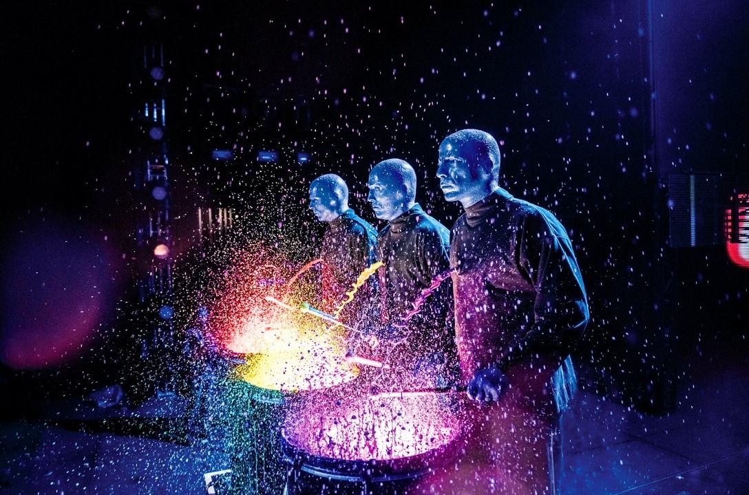 Three bald blue men dressed in black playing drums in front of a black background. Colored paint is splashing off the drums.