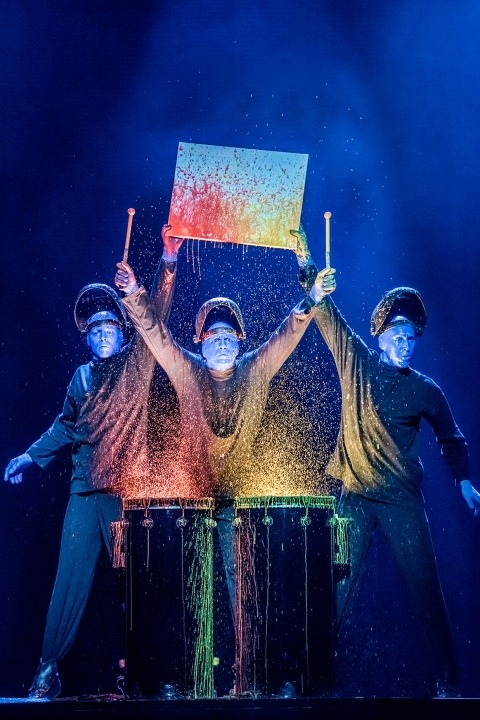 Three bald blue men dressed in black playing drums in front of a black background. Colored paint is splashing off the drums. Two of them are holding up a canvas with paint splattered on it.
