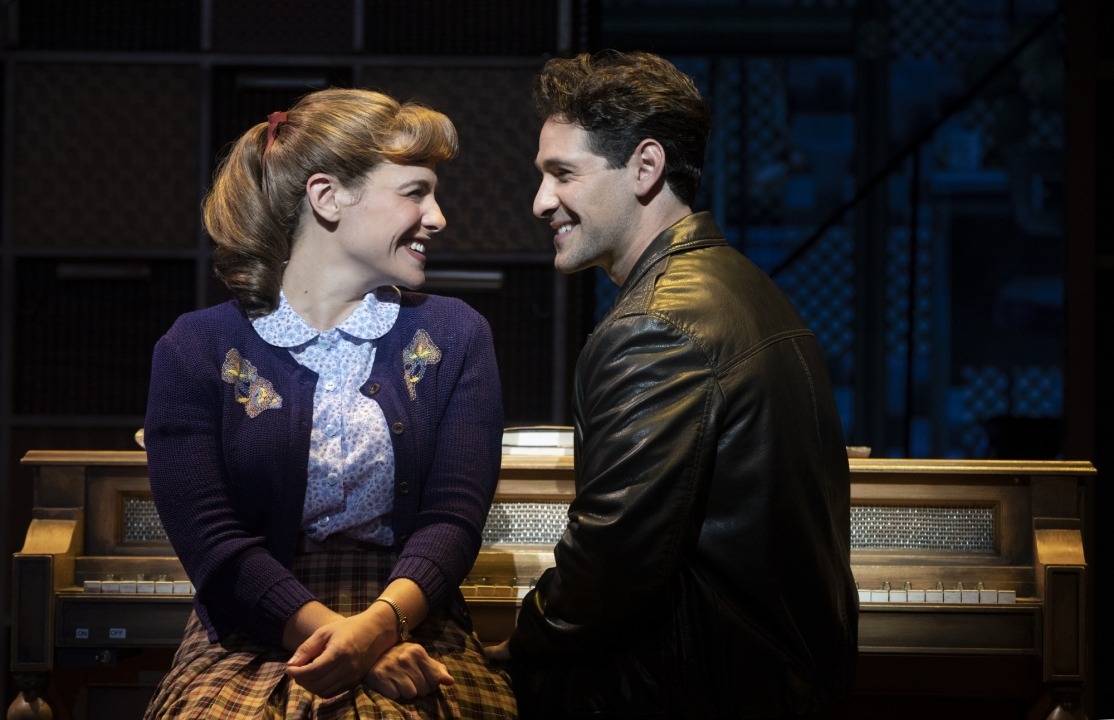 Man in a black leather jacket and woman in a purple cardigan sitting at a piano and smiling at each other