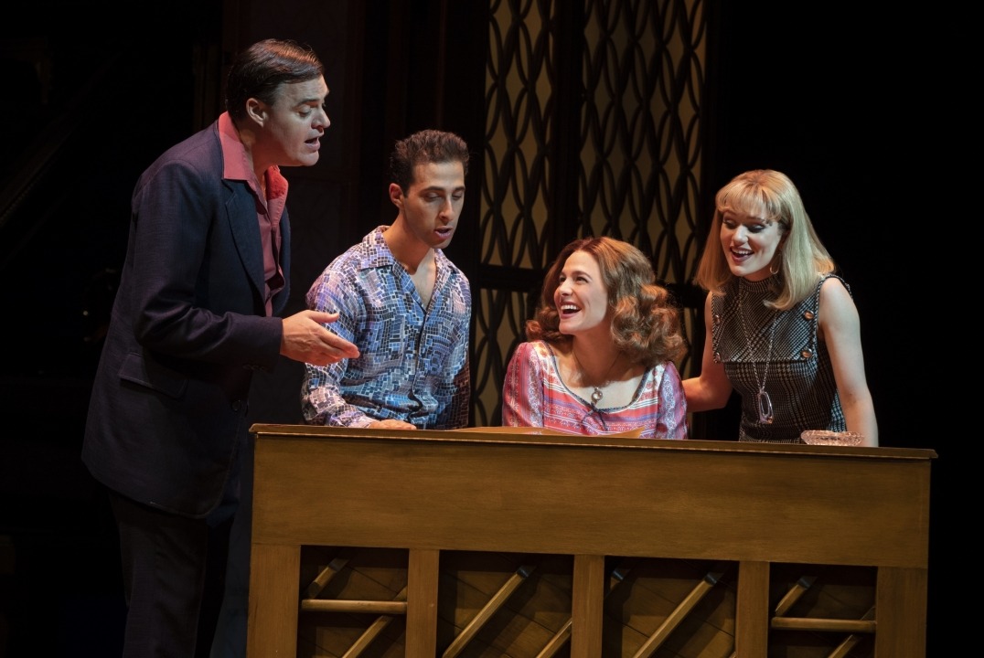 Two men and two women sitting behind a piano singing in front of a brown background