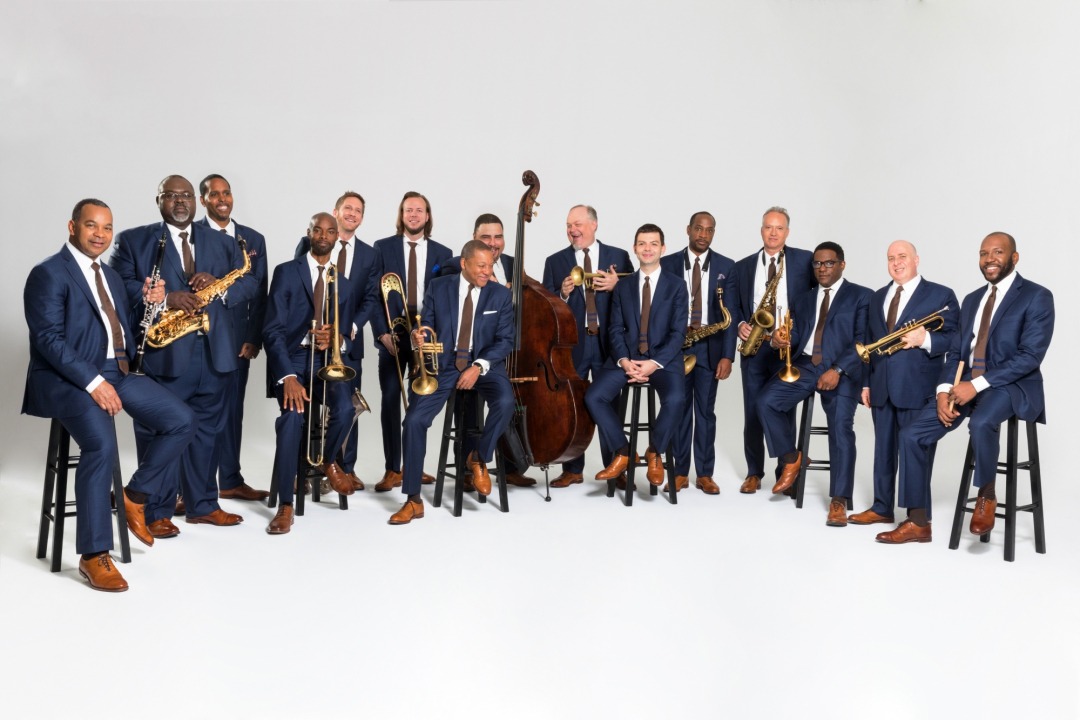 15 men, each wearing a blue suit, brown tie, and brown shoes, with a crisp white background. All men, except one, are holding a brass, wind, or percussion instrument. All of the men are smiling, some laughing.