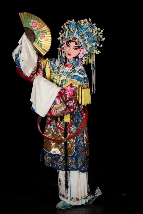 A Chinese woman wearing a headdress and robe made of ornate red, white, yellow, and blue fabric stands holding a gold fan with flowers on it at an angle near her face in front of a black background.