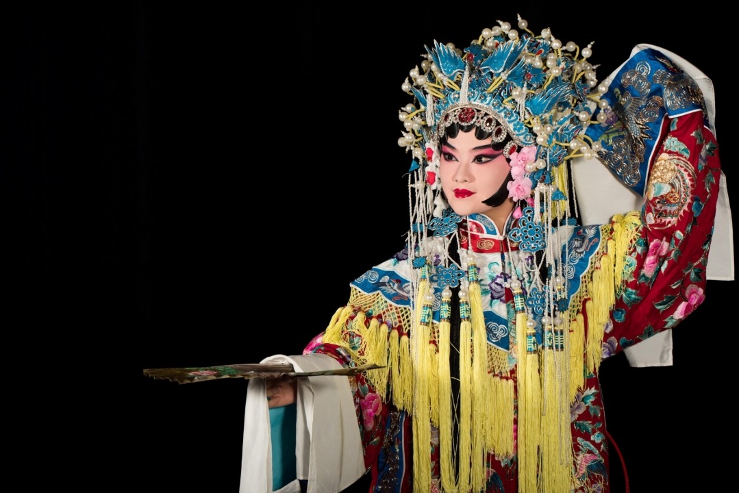 A Chinese woman wearing a headdress and robe made of ornate red, white, yellow, and blue fabric stands holding a gold fan parallel to the floor with her other hand up near her ear in front of a black background.