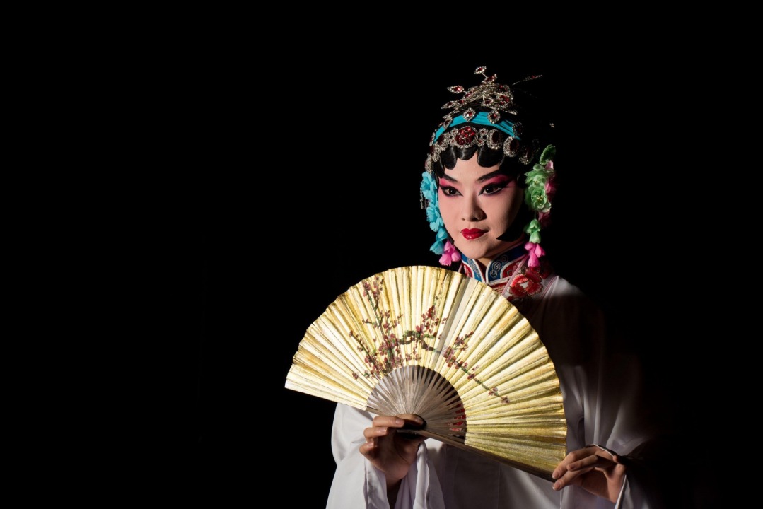 A Chinese woman dressed in white with a gold and blue headband stands holding a gold fan directly in front of her chest in front of a black background.