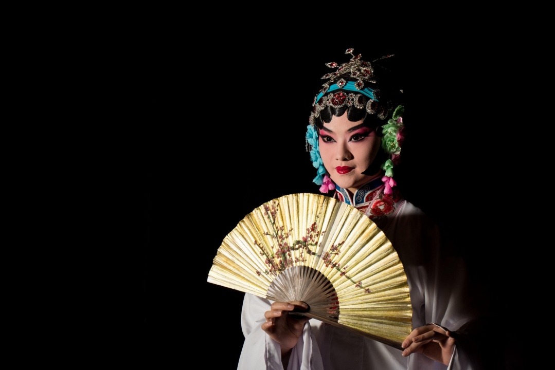 A Chinese woman dressed in white with a gold and blue headband stands holding a gold fan directly in front of her chest in front of a black background.